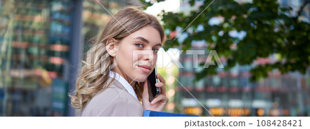 Close up portrait of corporate woman calling on mobile phone, walking on street with work documents in hand, wearing business suit 108428421