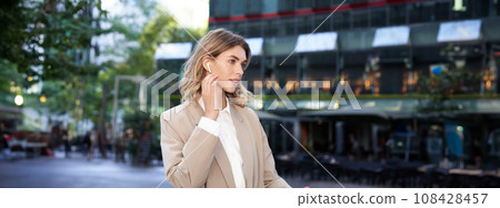 Stylish corporate woman in wireless headphones and suit, posing on street near office buildings 108428457