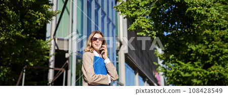 Working lady in sunglasses, saleswoman talking on mobile phone in front of office building, standing on street, holding folder 108428549