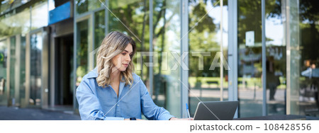 Successful businesswoman working outdoors, sitting on fresh air with laptop, documents and papers, looking at computer screen 108428556