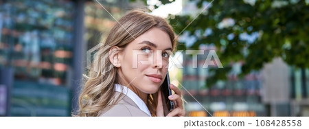 Close up shot of businesswoman talking on mobile phone. Corporate woman calling someone, looking around, standing outdoors Close up shot of businesswoman talking on mobile phone. Corporate woman calling someone, looking around, standing outdoors 108428558