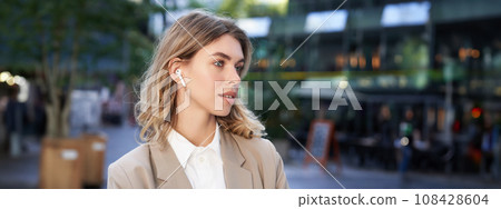 Close up portrait of blond businesswoman, confident corporate woman in headphones and beige suit, posing outdoors on street of city center Close up portrait of blond businesswoman, confident corporate woman in headphones and beige suit, posing outdoors on street of city center 108428604