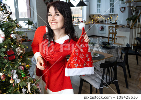 Selfie portrait of a woman in a red Santa dress in a home interior with a Christmas tree and New Year decor. Preparation for the holidays, party Selfie portrait of a woman in a red Santa dress in a home interior with a Christmas tree and New Year decor. Preparation for the holidays, party 108428610