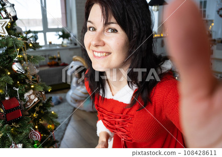 Selfie portrait of a woman in a red Santa dress in a home interior with a Christmas tree and New Year decor. Preparation for the holidays, party 108428611