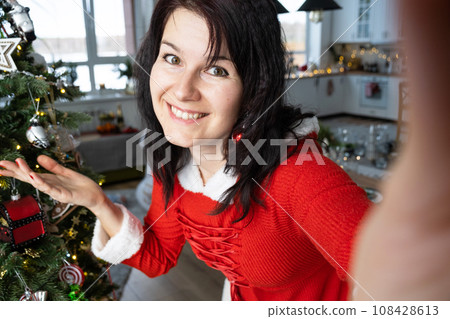 Selfie portrait of a woman in a red Santa dress in a home interior with a Christmas tree and New Year decor. Preparation for the holidays, party Selfie portrait of a woman in a red Santa dress in a home interior with a Christmas tree and New Year decor. Preparation for the holidays, party 108428613