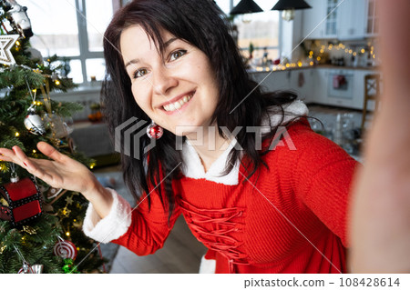 Selfie portrait of a woman in a red Santa dress in a home interior with a Christmas tree and New Year decor. Preparation for the holidays, party 108428614