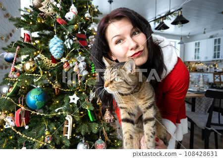 Selfie funny portrait of a woman in a red Santa dress and domestic cat in a home interior with a Christmas tree and New Year decor. Preparation for the holidays, party 108428631
