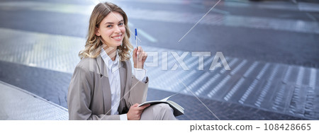 Business woman in suit sits in city centre, writes down, takes notes, holds pen and notebook, brainstorms, creates ideas 108428665