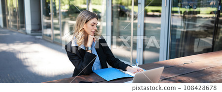Successful businesswoman working outdoors. Corporate woman sitting on bench with laptop, writing, taking notes during work meeting Successful businesswoman working outdoors. Corporate woman sitting on bench with laptop, writing, taking notes during work meeting 108428678