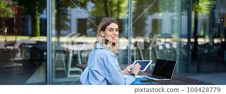 Portrait of successful businesswoman works outside on fresh air, sits near laptop, holds digital tablet and looks at camera 108428779