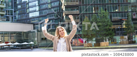 Corporate woman celebrating her victory outside on street. Happy businesswoman raising hands up and triumphing from excitement Corporate woman celebrating her victory outside on street. Happy businesswoman raising hands up and triumphing from excitement 108428800