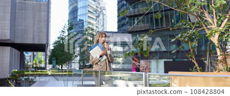 Corporate people. Beautiful business woman with laptop and documents, drinks her coffee in morning near office building, standing in city center Corporate people. Beautiful business woman with laptop and documents, drinks her coffee in morning near office building, standing in city center 108428804