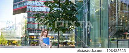 Vertical shot of young businesswoman in beige suit, holding documents in hands, looking confident at camera, standing outdoors in city 108428813