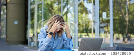 Portrait of young working woman sitting near laptop and feeling frustrated, disappointed. Businesswoman looks troubled while works on project, sits outdoors 108428999
