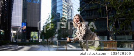 Happy smiling saleswoman stands on street on coffee break, talking on mobile phone and drinks takeaway drink Happy smiling saleswoman stands on street on coffee break, talking on mobile phone and drinks takeaway drink 108429109