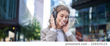 Portrait of smiling corporate woman, girl in suit listening music in headphones, sitting in city centre Portrait of smiling corporate woman, girl in suit listening music in headphones, sitting in city centre 108429124