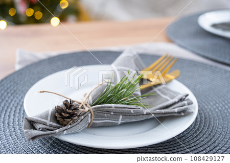 Serving a festive table with plates, forks, knives, napkins close-up tissue in a cage decorated for Christmas and New Year pine branch with a cone. Eco-style, minimalism Serving a festive table with plates, forks, knives, napkins close-up tissue in a cage decorated for Christmas and New Year pine branch with a cone. Eco-style, minimalism 108429127