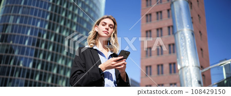 Low angle shot of young female manager, businesswoman in suit, holding mobile phone, standing with smartphone on street Low angle shot of young female manager, businesswoman in suit, holding mobile phone, standing with smartphone on street 108429150