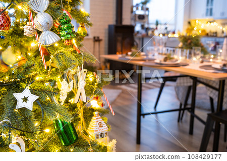 Festive interior of house is decorated for Christmas and New Year in loft style with black stove, fireplace, Christmas tree. Warm studio room with set table, burning wood, cozy and heating of home Festive interior of house is decorated for Christmas and New Year in loft style with black stove, fireplace, Christmas tree. Warm studio room with set table, burning wood, cozy and heating of home 108429177