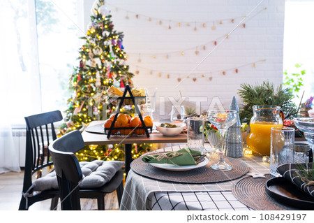 Festive interior of house is decorated for Christmas and New Year in loft style with black stove, fireplace, Christmas tree. Warm studio room with set table, burning wood, cozy and heating of home 108429197