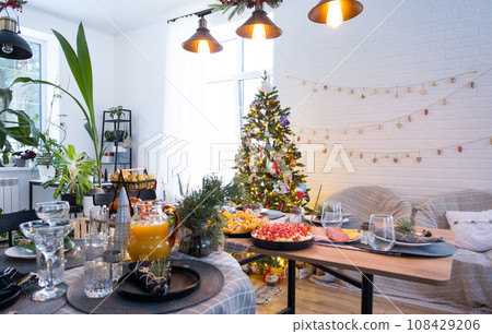 Festive interior of house is decorated for Christmas and New Year in loft style with black stove, fireplace, Christmas tree. Warm studio room with set table, burning wood, cozy and heating of home Festive interior of house is decorated for Christmas and New Year in loft style with black stove, fireplace, Christmas tree. Warm studio room with set table, burning wood, cozy and heating of home 108429206