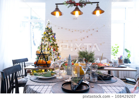 Festive interior of house is decorated for Christmas and New Year in loft style with black stove, fireplace, Christmas tree. Warm studio room with set table, burning wood, cozy and heating of home Festive interior of house is decorated for Christmas and New Year in loft style with black stove, fireplace, Christmas tree. Warm studio room with set table, burning wood, cozy and heating of home 108429207