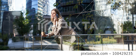 Stylish office woman in beige suit, talking on mobile phone while standing outside on street with morning coffee Stylish office woman in beige suit, talking on mobile phone while standing outside on street with morning coffee 108429235