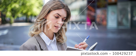 Woman working, writing down thoughts in her planner, sitting in city centre in business clothing Woman working, writing down thoughts in her planner, sitting in city centre in business clothing 108429237
