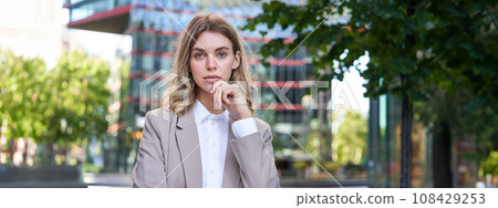 Portrait of confident corporate woman, young female ceo manager look at camera, posing in beige suit near office buildings, standing on street 108429253