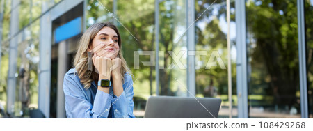 Dreamy woman sitting with laptop outdoors on street. Smiling businesswoman daydreaming, imaging smth while working outside office building 108429268