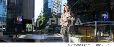 Stylish office woman in beige suit, talking on mobile phone while standing outside on street with morning coffee 108429269