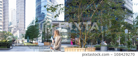 Vertical shot of smiling office girl in suit, drinks her coffee outdoors in city center, standing near office building, grabs takeaway 108429270
