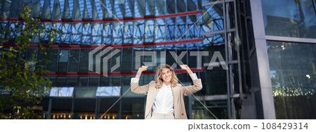 Happy businesswoman dancing on street, raising hands up. Corporate woman celebrates her victory or success 108429314