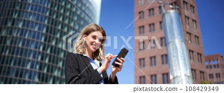 Low angle shot of businesswoman standing on street on sunny day, holding smartphone and using app, text message Low angle shot of businesswoman standing on street on sunny day, holding smartphone and using app, text message 108429349