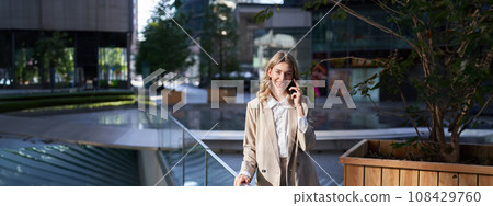 Successful businesswoman talking on mobile phone with confident smile, dealing with business calls, standing on street in city 108429760