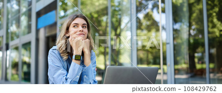 Portrait of young corporate woman working outdoors. Freelancer smiling, sitting with laptop outside. Businesswoman sitting outdoors with computer 108429762