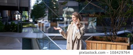 Beautiful young businesswoman in suit talks on mobile phone on street. Corporate woman calls someone on phone while stands outside 108429764
