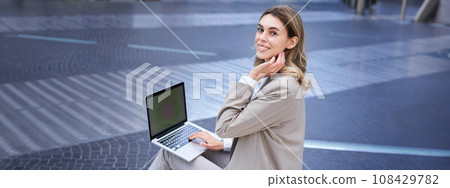 Young digital nomad, businesswoman sitting with laptop in city centre. Office worker using computer outdoors Young digital nomad, businesswoman sitting with laptop in city centre. Office worker using computer outdoors 108429782