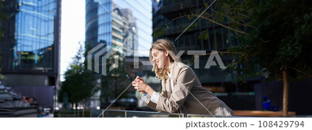 Smiling businesswoman using her mobile phone on a break, outside of office, looking at smartphone screen while messaging someone 108429794