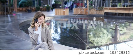 Vertical shot of corporate woman sitting outdoors with laptop, talking on mobile phone, working ouside office building while waiting for someone 108429815