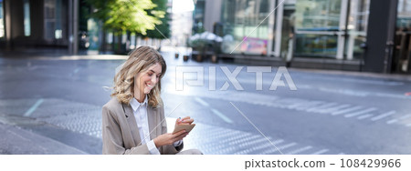 Businesswoman sitting with laptop, wearing suit, writing down notes in her planner, being in city centre outdoors 108429966