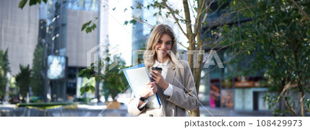 Smiling businesswoman standing on street, holding work documents, laptop and drinking takeaway coffee 108429973