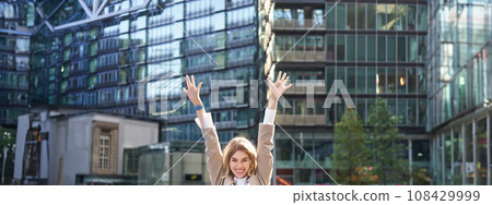 Corporate woman celebrating her victory outside on street. Happy businesswoman raising hands up and triumphing from excitement 108429999