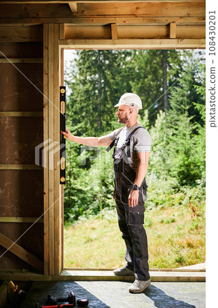 Carpenter building wooden-framed house near the forest. Bearded man inspects the walls for levelness using a spirit level. Concept of environmentally friendly construction. Carpenter building wooden-framed house near the forest. Bearded man inspects the walls for levelness using a spirit level. Concept of environmentally friendly construction. 108430201