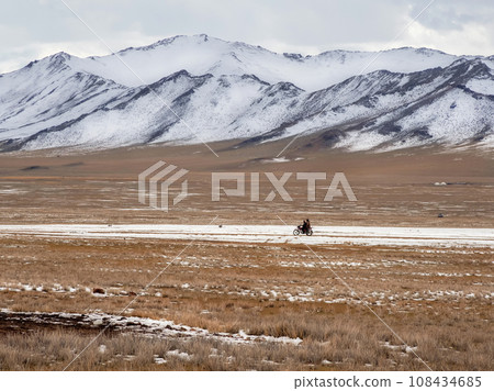 Beautiful Mongolian winter landscape snow mountain with a small figure of a ride on a black motorcycle through a wide snow-covered valley against the backdrop of high mountains. Beautiful Mongolian winter landscape snow mountain with a small figure of a ride on a black motorcycle through a wide snow-covered valley against the backdrop of high mountains. 108434685