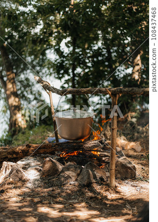 Travel a kettle over a fire burning on the river and sunset backgroun. Cooking over a campfire. Travel a kettle over a fire burning on the river and sunset backgroun. Cooking over a campfire. 108437468