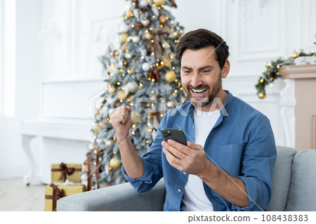 Man celebrating New Year and Christmas at home sitting on sofa, celebrating victory and successful news received on phone, using application on smartphone. 108438383