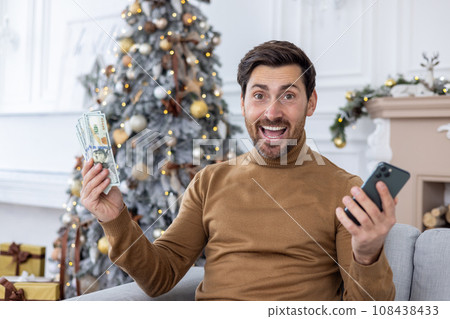 Portrait successful man sitting near the Christmas tree at home, smiling and looking at the camera, holding money, dollars, cash, and a phone, celebrating the successful results of the achievement. 108438433