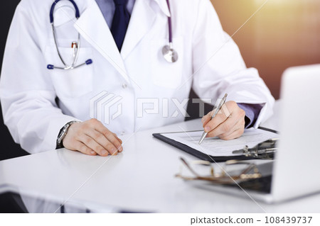 Unknown male doctor sitting and working with clipboard of medication history record in a darkened clinic, glare of light on the background, close-up of hands 108439737