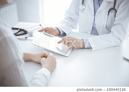 Doctor and patient sitting opposite each other at the desk in clinic. The focus is on female physician's hands pointing into tablet computer touchpad, close up. Medicine concept 108440186